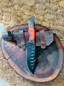 The Texan Knives Hunting Skinning Knife features a 10" J2 steel blade and a wangi/resin handle, resting on its leather sheath atop a round wooden slab with rustic texture and natural patterns.