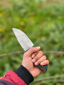 A hand in a red and black jacket holds the Texan Knives 10" Handmade Damascus Steel Hunting Knife with a rosewood handle from their Gutting Knives line, shown against a blurred green outdoor background.