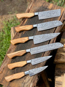 Six Texan Knives Sharp Kitchen Knife Sets, each featuring a 13" Damascus steel blade, olivewood handle, and leather sheath, are arranged side by side outdoors on rustic wood with grass in the background.