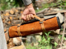 A person holds a roll-up leather and canvas tool bag with tan straps, outdoors among logs—ideal for carrying the Texan Knives Sharp Kitchen Knife Set featuring a 13" Damascus steel blade, olivewood handle, and leather sheath.