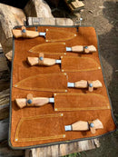 The Texan Knives Sharp Kitchen Knife Set features a 13" Damascus steel blade, olivewood handle, and comes in a brown leather sheath, with each knife displayed outdoors on stacked logs.