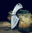 The Texan Knives Handmade Damascus Steel Fixed Blade Pocket Knife – Full Tang Skinner with a curved handle and intricate swirling designs leans against a large rock, set on wood with a dark background. Comes with a leather sheath.