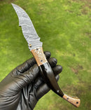 A hand in a black glove holds the Texan Knives Custom Handmade Damascus Blade EDC Folding Pocket Knife with a color bone handle and brass engraved bolster, set against blurred green grass; leather sheath included.