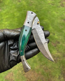 A black-gloved hand holds the Texan Knives Custom Handmade Damascus Blade EDC Folding Pocket Knife with a 3-inch blade, color bone handle, and brass engraved bolster; grassy outdoors form the background.