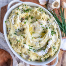 A white oval dish of creamy mashed potatoes, topped with chives, butter swirls, and black pepper. Garlic and SALTOPIA Infused Sea Salts' BEN THERE, D'ONION THAT onion salt are visible in the background.
