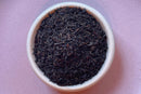 A small white bowl filled with loose World Spice Earl Grey black tea leaves, infused with bergamot oil for a classic aroma, is placed on a light purple surface.