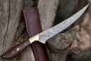 The Texan Knives Damascus Fillet Knife – 13" features a patterned blade, rosewood handle, and brass bolster, shown on a tree trunk beside its brown leather sheath.