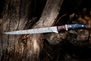 A Handmade Damascus Fillet Knife 13” by Texan Knives, featuring a patterned blade and a Buffalo Horn, Rosewood & Brass handle with leather sheath, rests on rugged tree bark outdoors.