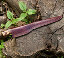 The Texan Knives Handmade Damascus Fillet Knife 8” with a rosewood & olive wood handle and leather sheath rests on a weathered tree stump, surrounded by green leaves and plants.