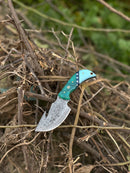 The Texan Knives Elite Custom Handmade Damascus Steel Guthook Skinning Knife, 7.5" with a resin handle and leather sheath, lies among dry branches outdoors, green foliage blurred in the background.