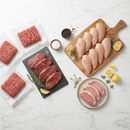 An assortment from the Dude Food Essentials Meat Box by Dude Food, featuring raw ground beef patties, flat iron steak slices with rosemary and lemon, boneless chicken breasts seasoned with herbs and lemon on a cutting board, and pork chops on a plate—all presented on a white surface.