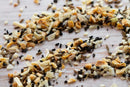 A close-up of the World Spice Americana Tasting Flight by World Spice showcases a vibrant blend of dried spices and seeds—like sesame seeds, dried onion, dried garlic, and poppy seeds—spread across a wooden surface, perfect for exploring aromatic spice blends inspired by regional flavors from across the USA.