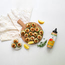 A round wooden board and small plate with cucumber bites topped with salmon and herbs, a bottle of Terrapin Ridge Farms Avo Everything Aioli Squeeze, lemon wedges, fresh dill, and a white napkin on a white surface.