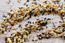 A close-up of World Spice Everything Bagel Spice by World Spice—a scattered mix of dried onions, garlic, sesame seeds, poppy seeds, nigella seed, and other seasonings—spread enticingly across a wooden surface.