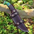 The Texan Knives Custom Handmade Damascus Skinning Knife features a 4.5" resin handle and comes with a brown leather sheath, shown resting on a tree branch with green foliage in the background—perfect for seasonal hunts.