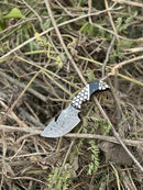 The Texan Knives Exquisite Custom Handmade Damascus Steel Guthook Skinning Knife features a 7.5” blade, unique resin handle, and comes with a leather sheath—shown outdoors on dry twigs and green plants.