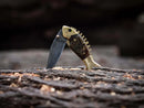 The Texan Knives Custom Handmade Fish Style Damascus Steel Folding Knife, featuring a sheep horn and brass handle, stands partially open in a textured wood piece with shavings. Includes leather sheath.