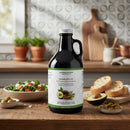 A bottle of Georgetown Olive Oil Co 50% Avocado Olive Oil + 50% Extra Virgin | High Polyphenols sits on a wooden kitchen counter next to sliced bread, a halved avocado, a fresh salad, and a bowl of green olives. Crafted by Georgetown Olive Oil Co., this oil is rich in polyphenols for added health benefits, making it an ideal choice for any wholesome kitchen. In the background, cutting boards and fresh herbs complete the inviting scene.