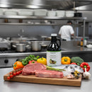 A cutting board with raw steak, fish, vegetables, and a bottle of Georgetown Olive Oil Co. 50% Avocado Olive Oil + 50% Extra Virgin | High Polyphenols sits in a kitchen. Pots, plates, and a person are visible in the background.