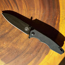 The Spaceman Knives Naiad EDC Pocket Knife by Spaceman Knives, featuring a textured black handle and titanium-plated blade, is open and rests on sunlit wood with warm, swirling grain patterns.