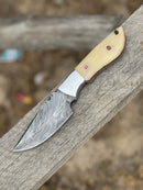 The Texan Knives Handmade 9" Damascus Steel Skinning Knife, featuring a camel bone handle with steel bolster and leather sheath, is displayed on a weathered wooden plank.