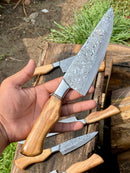 A hand holds a Texan Knives Sharp Kitchen Knife from the set, featuring a 13" Damascus steel blade and olivewood handle. Three similar knives with olivewood handles rest outdoors on rustic wood, framed by grass and soil.