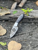 The FIXED BLADE POCKET KNIFE by Texan Knives, a personalized Damascus steel hunting knife with a patterned blade and dark handle, lies on weathered wood amid dry brown and green leaves on a rough gray surface.