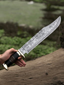 A hand holds the Texan Knives Authentic Crocodile Dundee Knife—16” handmade Damascus steel blade with buffalo horn or leather handle, brass guard, and leather sheath—set against a blurred outdoor background of greenery and a fallen log.