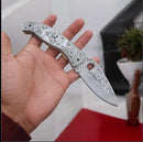 A person holds the Texan Knives Hand Engraved J2 Steel Pocket Knife – a 7.5" folding EDC with a detailed skull design on the handle. The background shows a plant and geometric red and white surfaces.