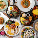 An overhead view of a table with assorted breakfast and brunch dishes, featuring eggs, pancakes, fruit, and an Everything Bagel—all enhanced with SALTOPIA Infused Sea Salts from the Saltopia Gift Set Exclusively for Foodie Fans: FOODIE AFICIONADO.