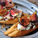 Toasted baguette slices with creamy cheese, prosciutto, fresh figs, and balsamic glaze, finished with SALTOPIA Infused Sea Salts from the FOODIE AFICIONADO Saltopia Gift Set for Foodie Fans, served on a silver plate.