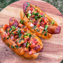 Two grilled hot dogs in buns topped with diced tomatoes, onions, jalapeños, cilantro, and a drizzle of Terrapin Ridge Farms Spicy Chipotle Garnishing Squeeze from Terrapin Ridge Farms, served on a wooden board.