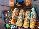 Four grilled corn cobs topped with cheese, chili powder, herbs, and Terrapin Ridge Farms Chile Lime Aioli Squeeze Garnishing Sauce are served with lime wedges. Terrapin Ridge Farms sauce bottles are visible in the background.