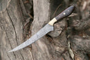 The Texan Knives Damascus Fillet Knife – 13" with a rosewood handle and leather sheath is displayed outdoors on a textured tree trunk.