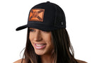 A smiling woman wears the Haka Hat Florida Flag Trucker Hat Leather in black, featuring a brown circular emblem with a geometric design on the front patch. The background is white.