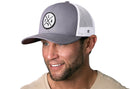 A smiling man with stubble wears the Haka Hat Florida Trucker Hat Gray-White FLA X Snapback, featuring a circular black logo. He pairs it with a light beige shirt and faces slightly to the right.