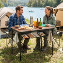 Two people enjoy snacks at the lake on a sunny day using the DirectDeals Folding Camping Table with Adjustable Heights Storage Net & Handle, with drinks and a thermos nearby and boats in the background.