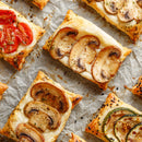 Rectangular puff pastry tarts with mushrooms, tomatoes, and zucchini are arranged on parchment paper. They're garnished with herbs and SALTOPIA Infused Sea Salts’ Saltopia Everything Bagel Salt for extra flavor.