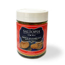 A jar of SALTOPIA Infused Sea Salts features the French Mustard Salt: MUSTARD BEEN LOVE, a savory Dijon mustard salt with a red label showing mustard and seeds—perfect for seasoning proteins.