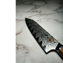 A close-up of the Spaceman Knives Interstellar Petty Chef Knife shows its patterned Damascus steel blade and wooden handle resting on light marble; the intricate wavy designs from Spaceman Knives are clearly visible.
