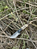 A 9" Damascus steel blade hunting knife by Texan Knives, featuring a sheep horn and resin handle with leather sheath, rests on dry twigs and leaves outdoors.