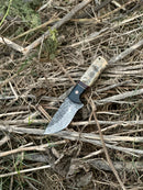 The Texan Knives Hunting Knife features a 9" Damascus steel blade and a polished sheep horn/resin handle, shown outdoors on dry twigs and grass. Comes with a leather sheath.