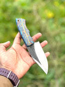 A hand holds the Texan Knives Good Hunting Knife, featuring a 9" handmade J2 steel blade, a blue and gold epoxy resin handle, and a leather sheath, set against a blurred green background.