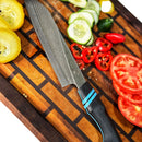 The Spaceman Knives Calypso Chef Knife features a patterned VG10 steel blade and a black handle with blue stripes, shown on a wooden cutting board with sliced squash, cucumber, chili peppers, and tomato—ideal for precise cuts.