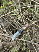 The Texan Knives Hunting Knife for Sale features a 9" Damascus steel blade and a sheep horn/resin handle with a leather sheath, displayed outdoors among dry twigs and branches.