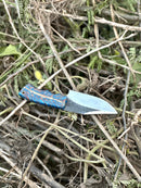 The Texan Knives Good Hunting Knife features a 9" handmade J2 steel blade and a patterned blue-orange epoxy resin handle, resting on dry branches and green foliage. Includes leather sheath.