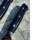 A close-up of two Spaceman Knives Interstellar Series steak knives featuring blue hexagonal patterns, metallic accents, and a round golden detail, resting on a white marble surface with shadows.
