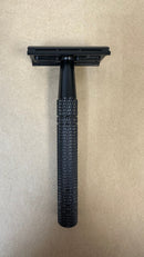 A Safety Razor by The Immaculate Beard, featuring a black metal design and a textured handle, stands vertically on a plain beige surface—poised to provide a close, precise shave when used with quality safety razor blades.