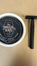 A black metal Safety Razor by The Immaculate Beard and a round container of The Immaculate Beard Twilight Shave Soap rest on a tan surface. The soap lid features details about its scent and weight, while the Safety Razor is designed to provide a close, comfortable shave.