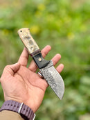 A hand holds the Texan Knives Hunting Knife—9" Damascus steel blade with a sheep horn/resin handle and leather sheath—against a blurred green outdoor background.
